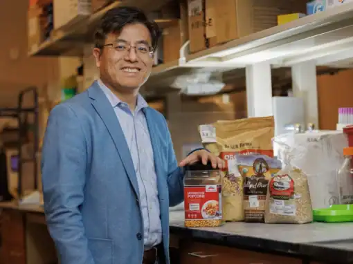 A smiling man in a blue blazer stands in a lab setting, holding a jar of mushroom popcorn. Several bags of grains and ingredients are displayed on the counter behind him.