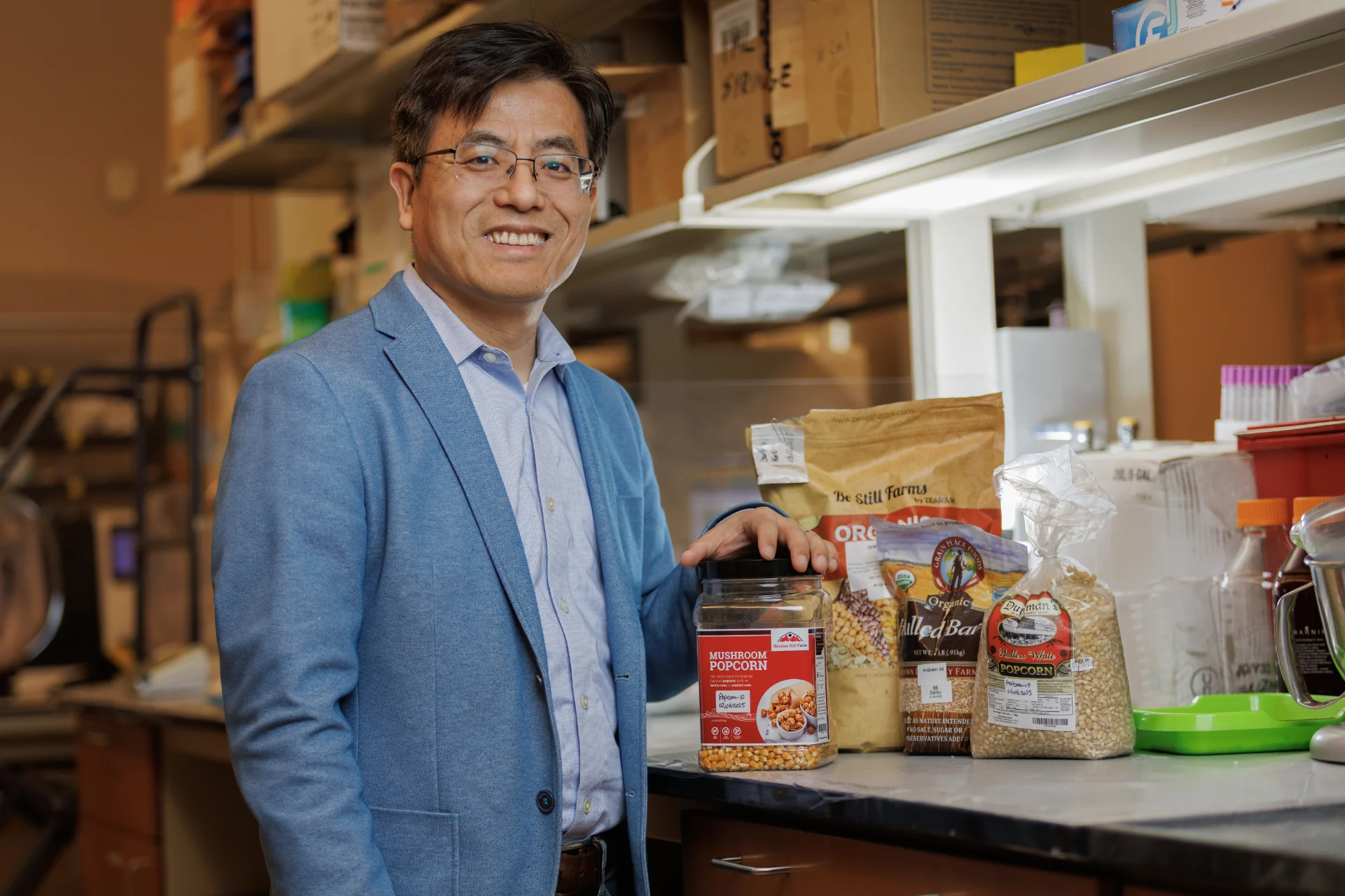 01_15_2026_Dr. Sang at NCRC 002 (1) A smiling man in a blue blazer stands in a lab setting, holding a jar of mushroom popcorn. Several bags of grains and ingredients are displayed on the counter behind him.