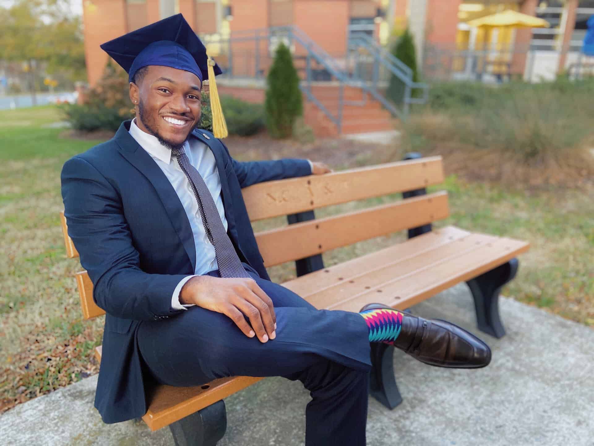 Graduation cap and gown worn by smiling young man sitting on park bench outdoors.