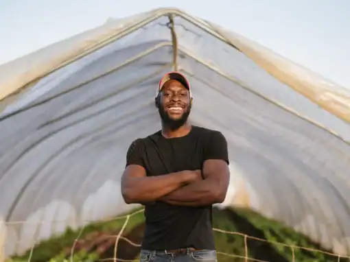 A smiling man stands with arms crossed inside a greenhouse, surrounded by rows of plants. He wears a black t-shirt and a cap, posing confidently against a backdrop of a clear sky.