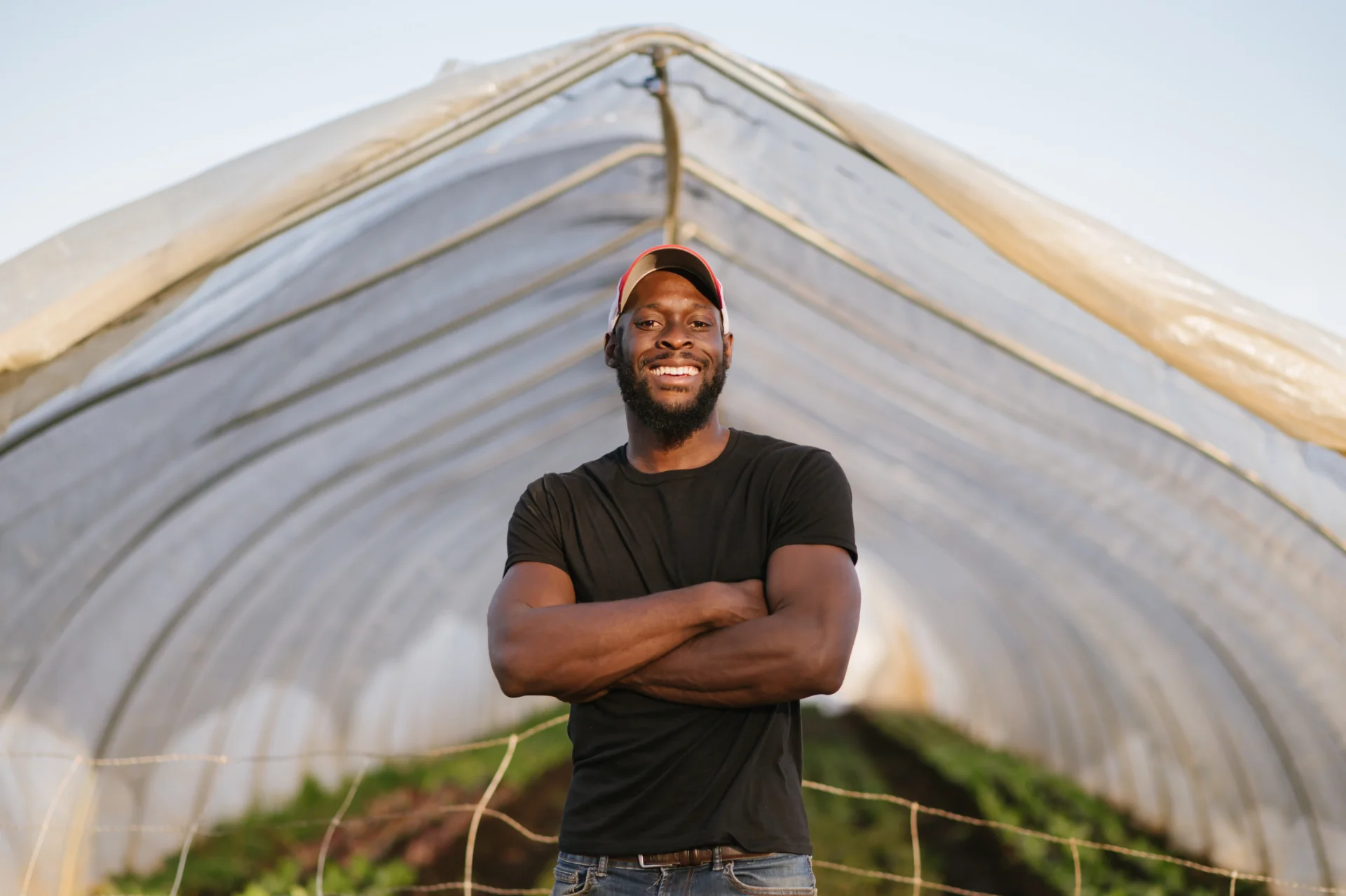 A smiling man stands with arms crossed inside a greenhouse, surrounded by rows of plants. He wears a black t-shirt and a cap, posing confidently against a backdrop of a clear sky.