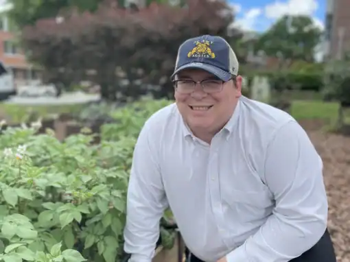 Smiling man in garden with plants, trees, and blue sky.