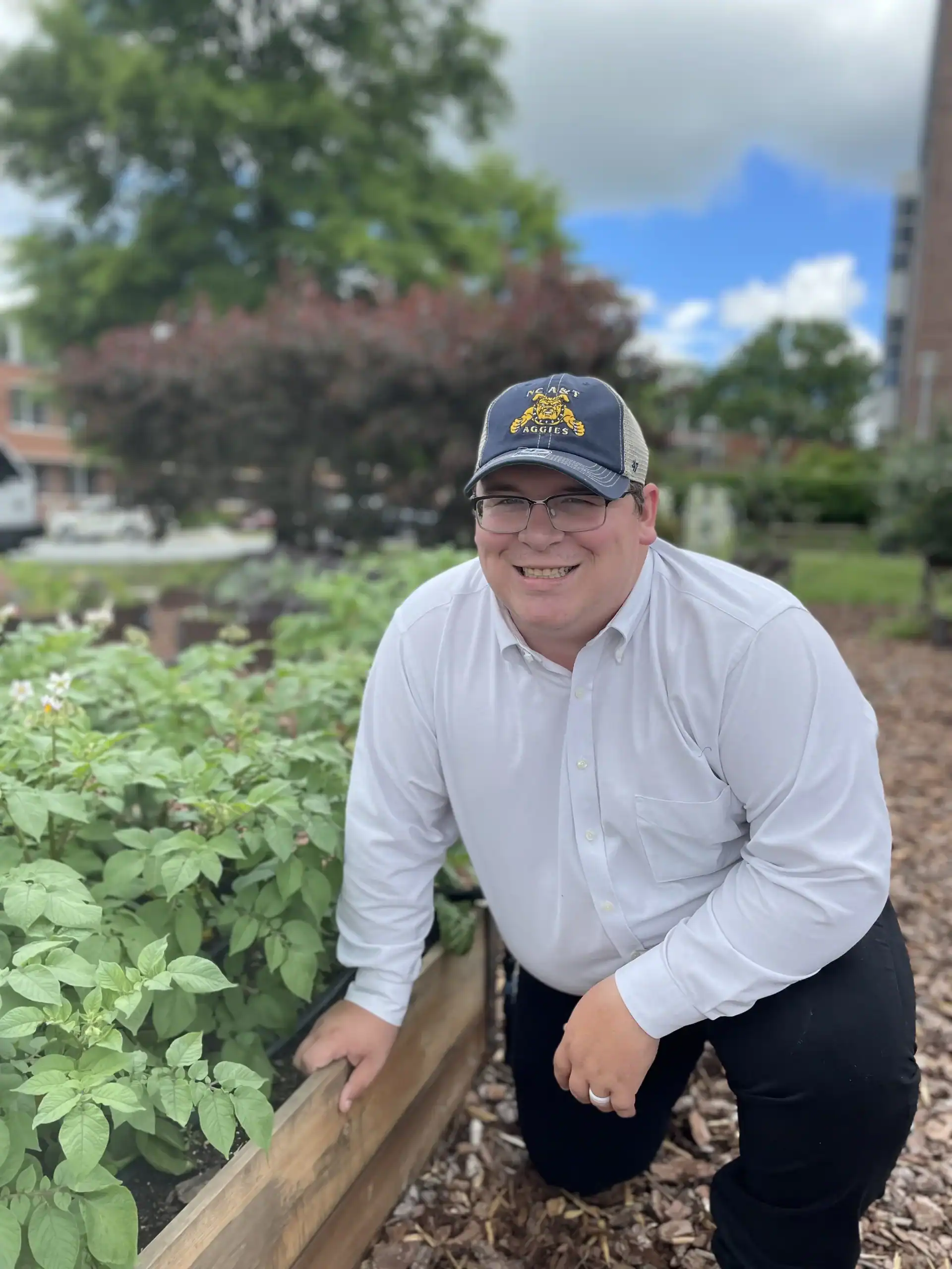 Smiling man in garden with plants, trees, and blue sky.