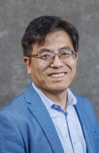Professional portrait of a smiling man in a blue suit and glasses.