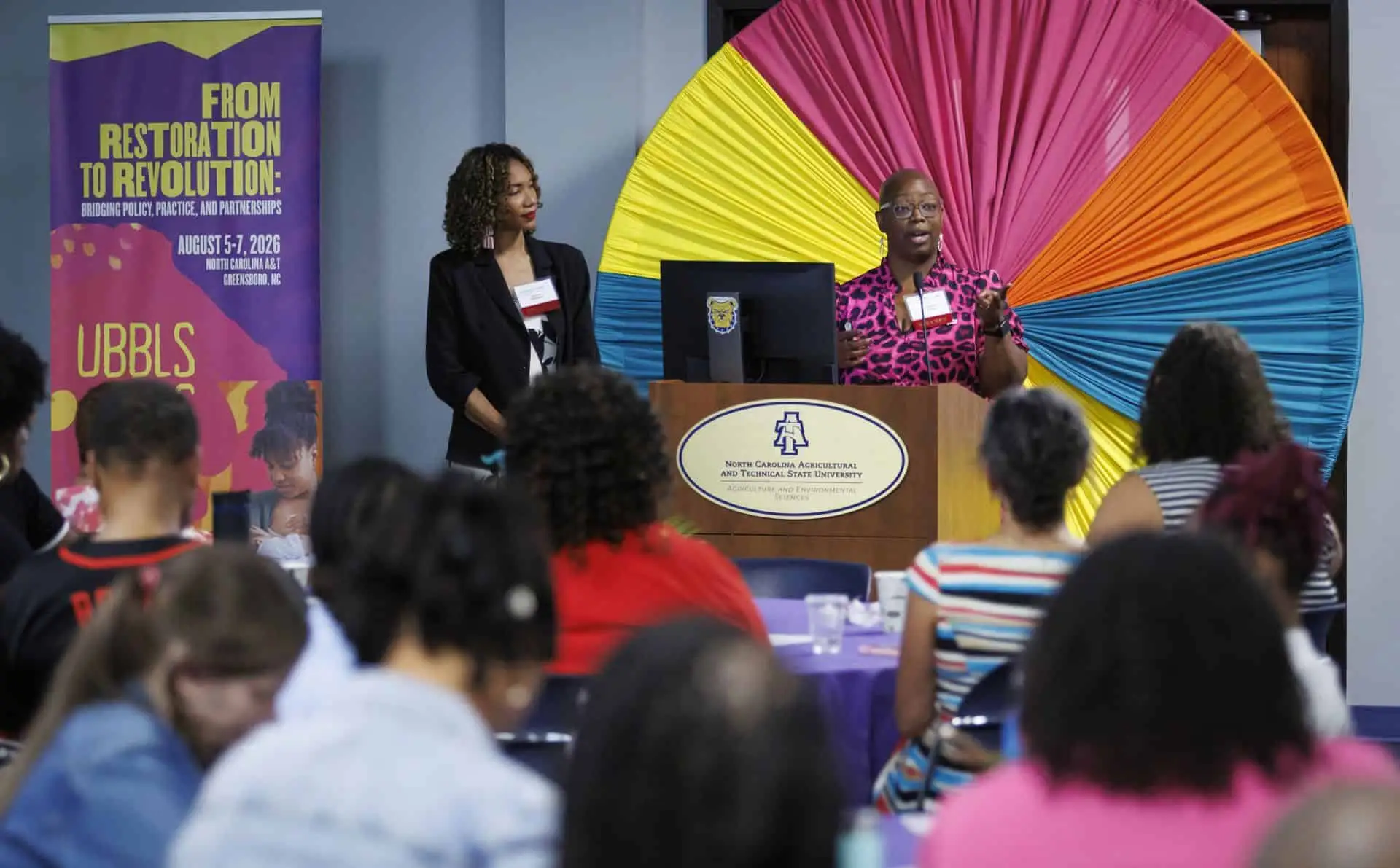 Diverse speakers at a conference with colorful backdrop and audience.