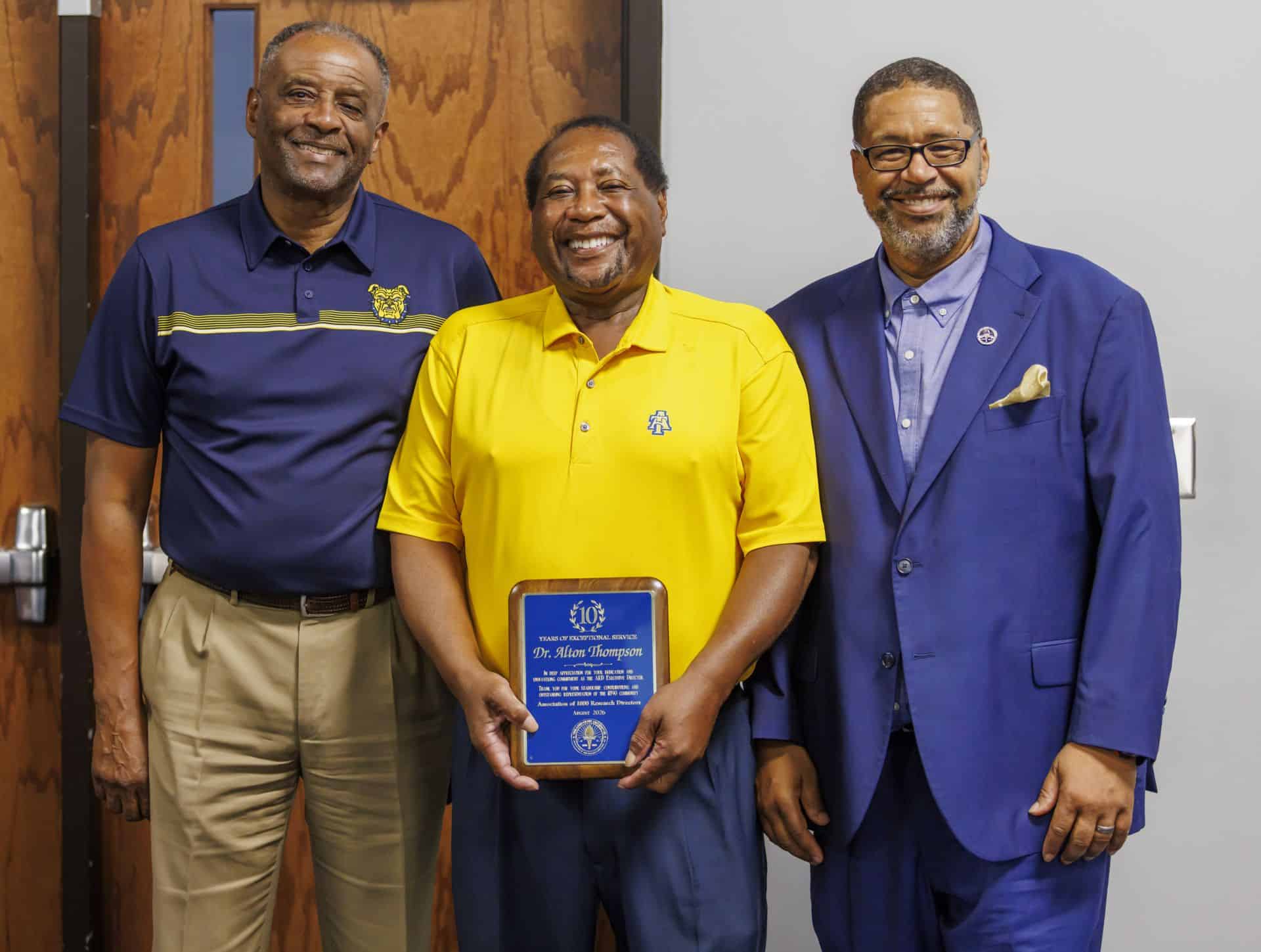 Three men at an award ceremony, one holding a plaque, smiling for the camera.