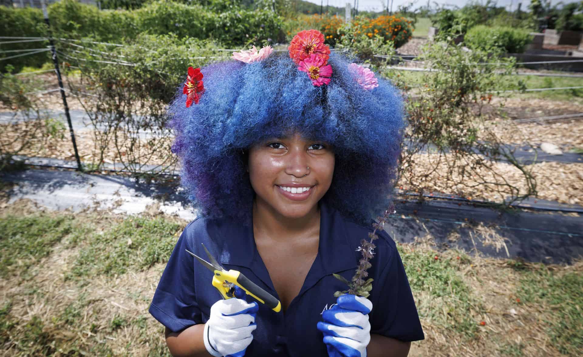 Young woman with vibrant blue and purple hair holding gardening tools outdoors.