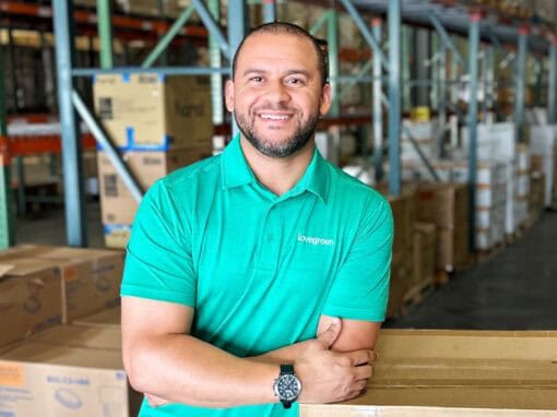 Smiling warehouse worker in teal polo shirt in storage facility with boxes and shelves.
