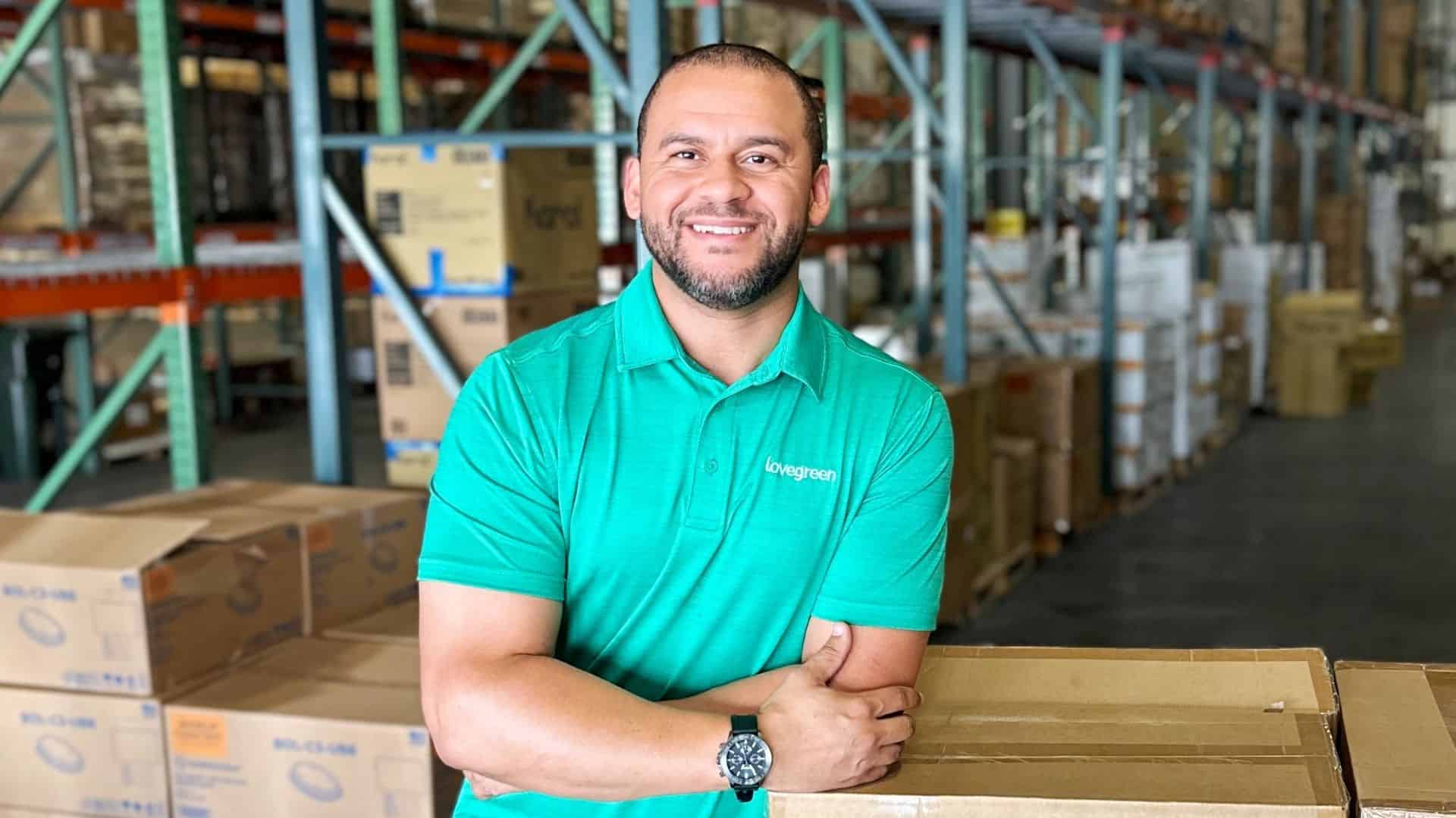 Smiling warehouse worker in teal polo shirt in storage facility with boxes and shelves.