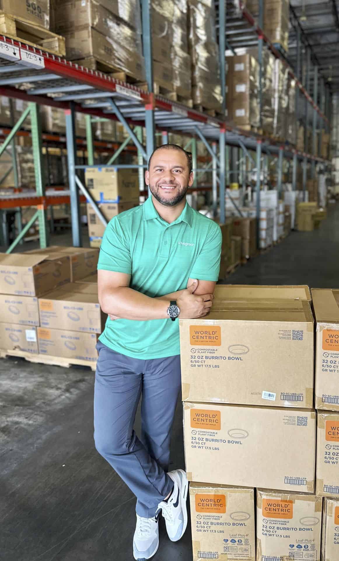 Smiling man in warehouse with stacked boxes and shelves in background.