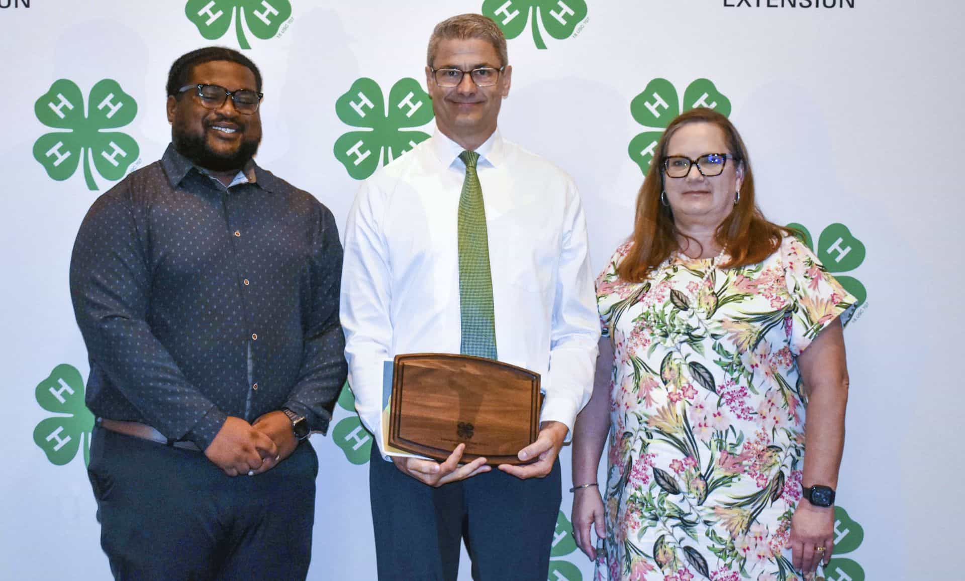 Three professionals at CAES News event with green four-leaf clover logos in background.
