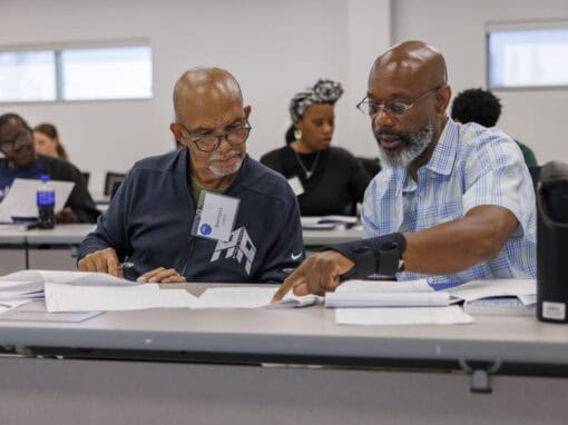 Two men collaborate over documents at a workshop or conference table.