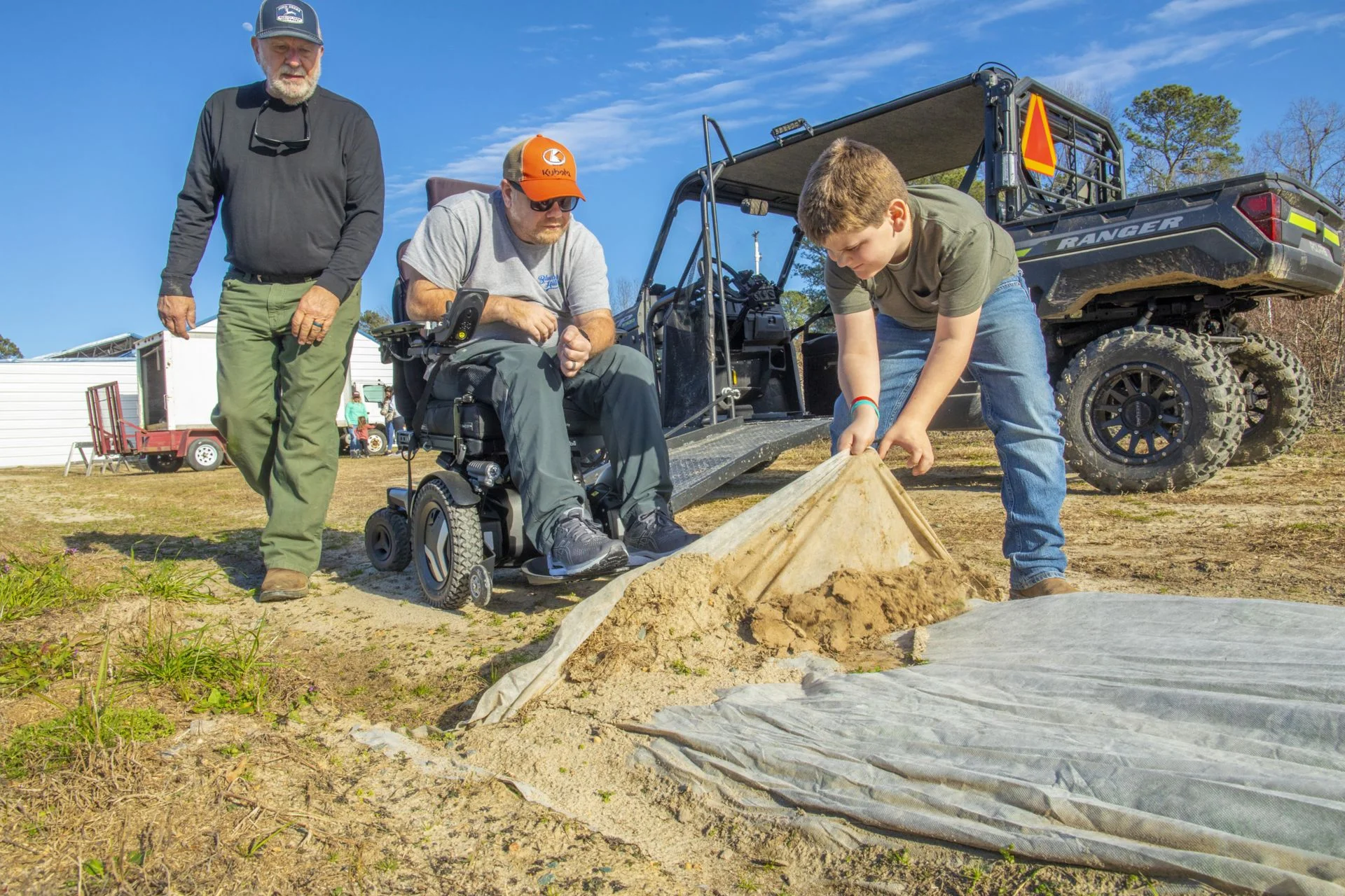 AgrAbility David Dyson 2343 – resized Three men work beside an off-road utility vehicle as one pulls back a tarp covering a mound of sand.